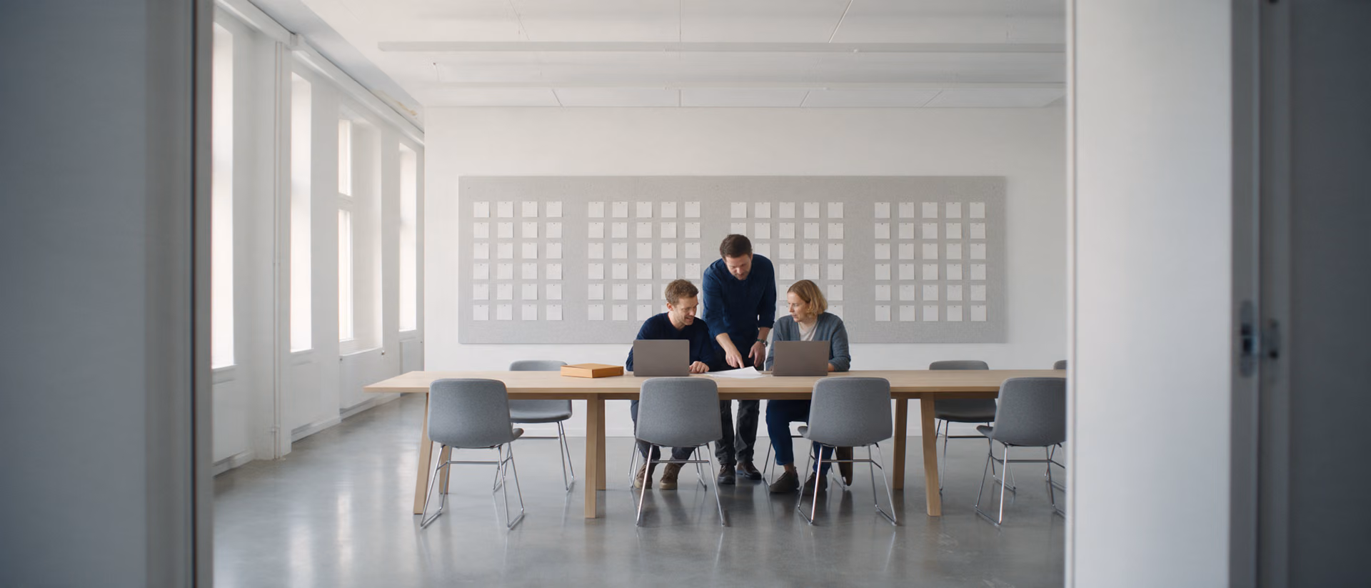 Three colleagues work at a long wooden table in a bright project room, a grid of blank cards pinned to the wall behind them.