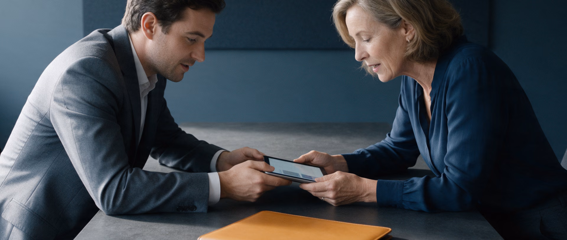 A man passes a tablet across a table to a woman in a meeting room, both leaning over it, a leather folder on the table between them.
