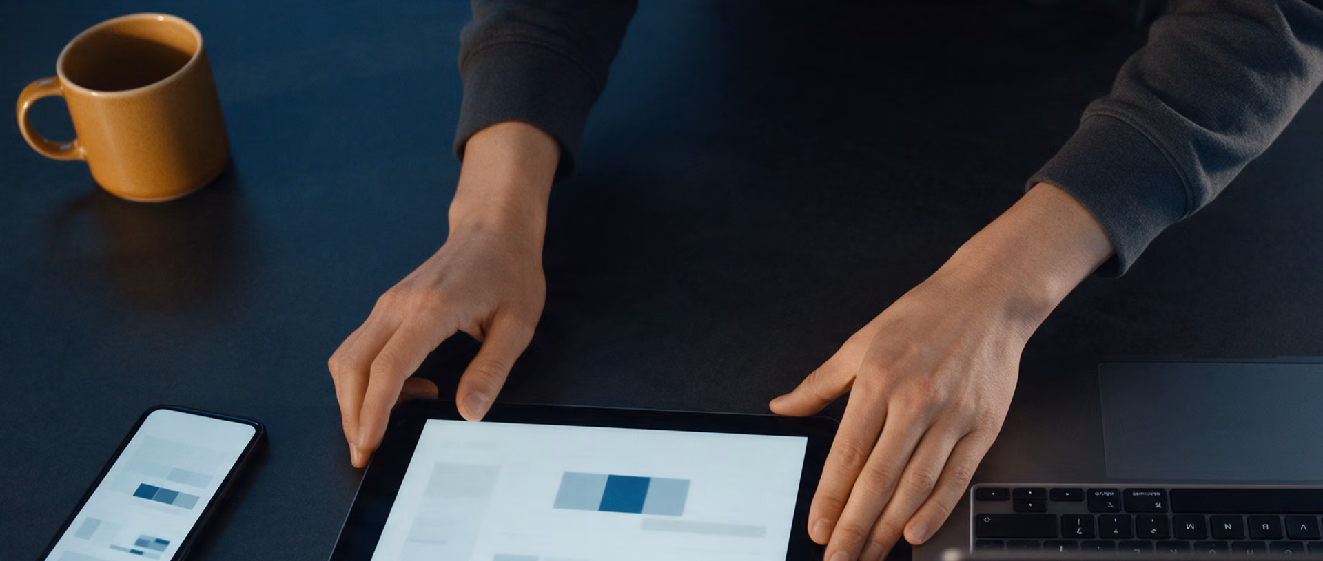 Two hands shift a tablet closer to a laptop on a dark table, a phone and a mug beside them, each screen showing blurred blocks.