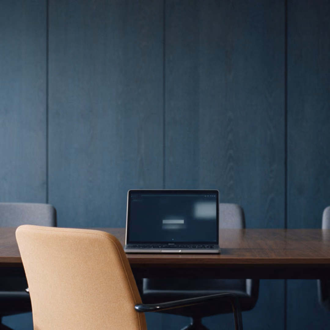 An empty boardroom with a dark wood-panelled wall, an open laptop on the table and one amber upholstered chair in the foreground.