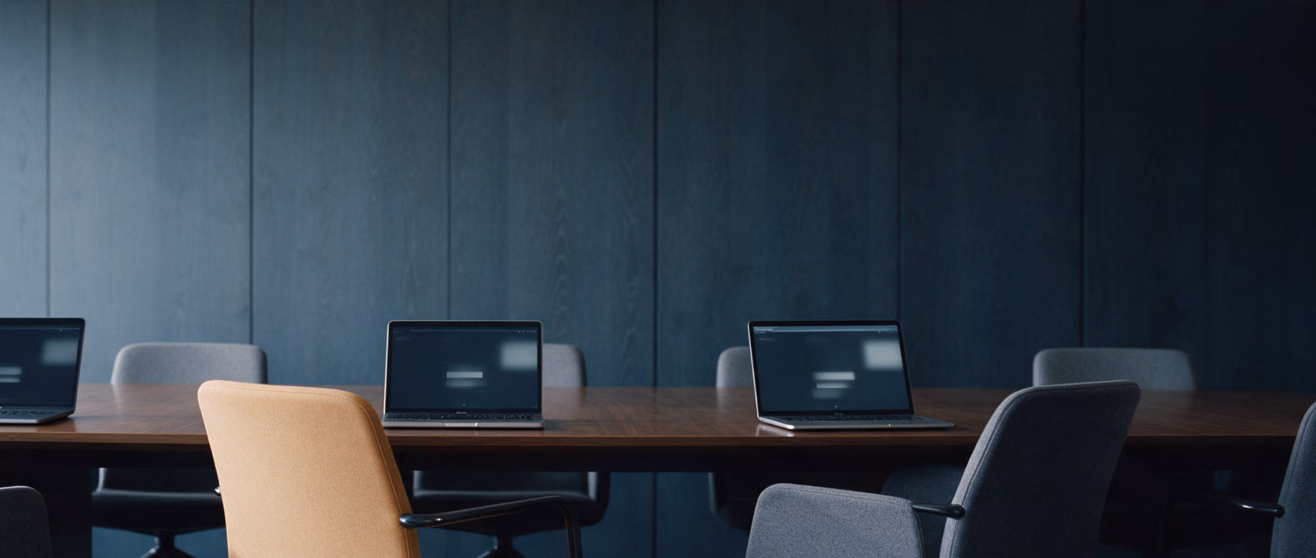 An empty boardroom with three open laptops spaced along a long dark table, their chairs pushed back in different directions.