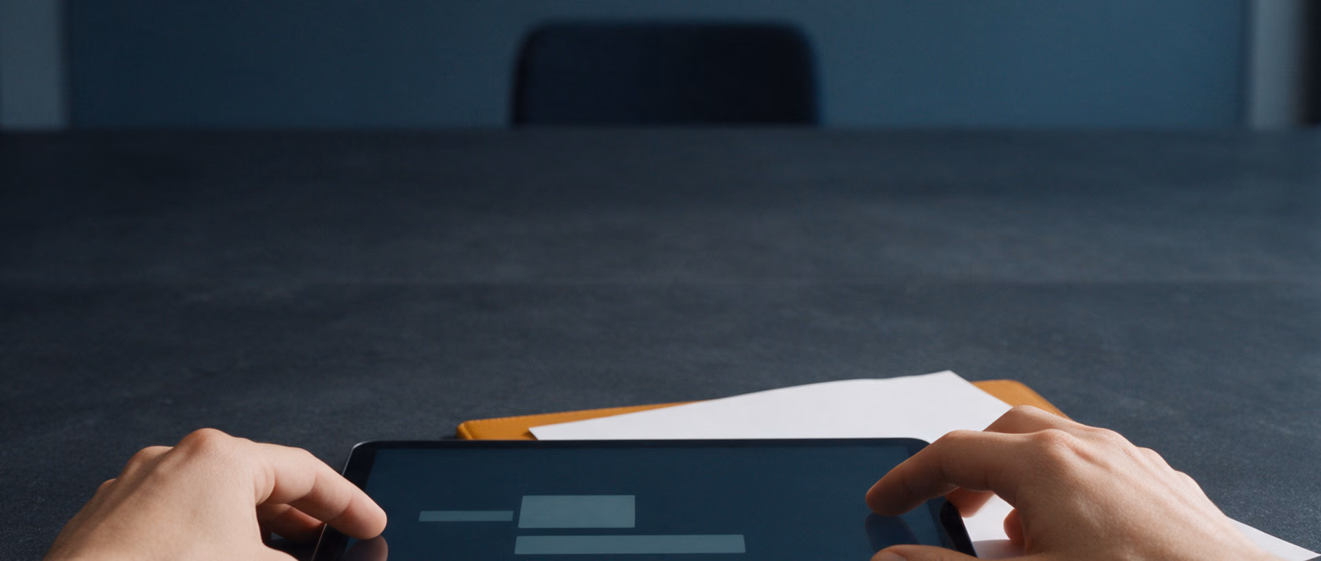 Two hands rest at the edges of a tablet lying flat on a dark meeting table after dark, next to a blank sheet of paper on a leather folder.