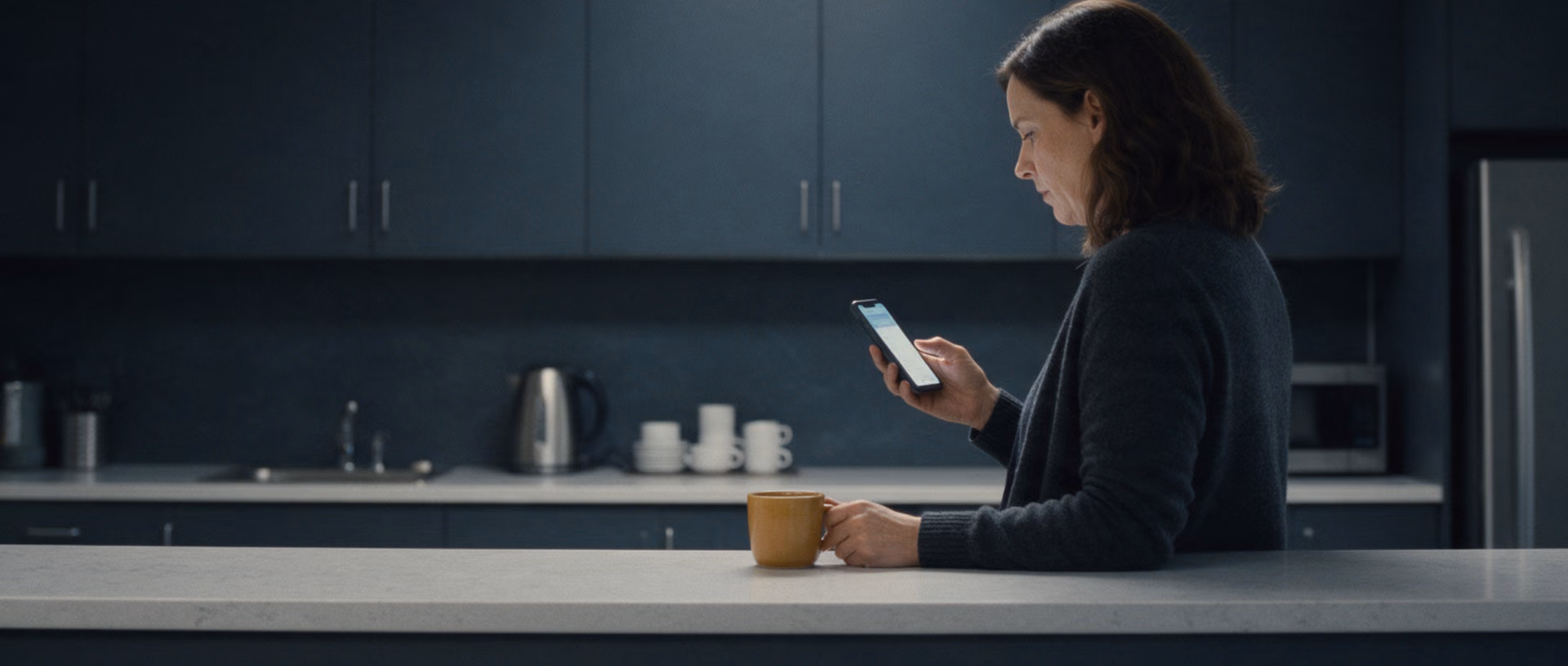 A woman stands at an office kitchen counter in the evening reading on her phone, a mug beside her hand.