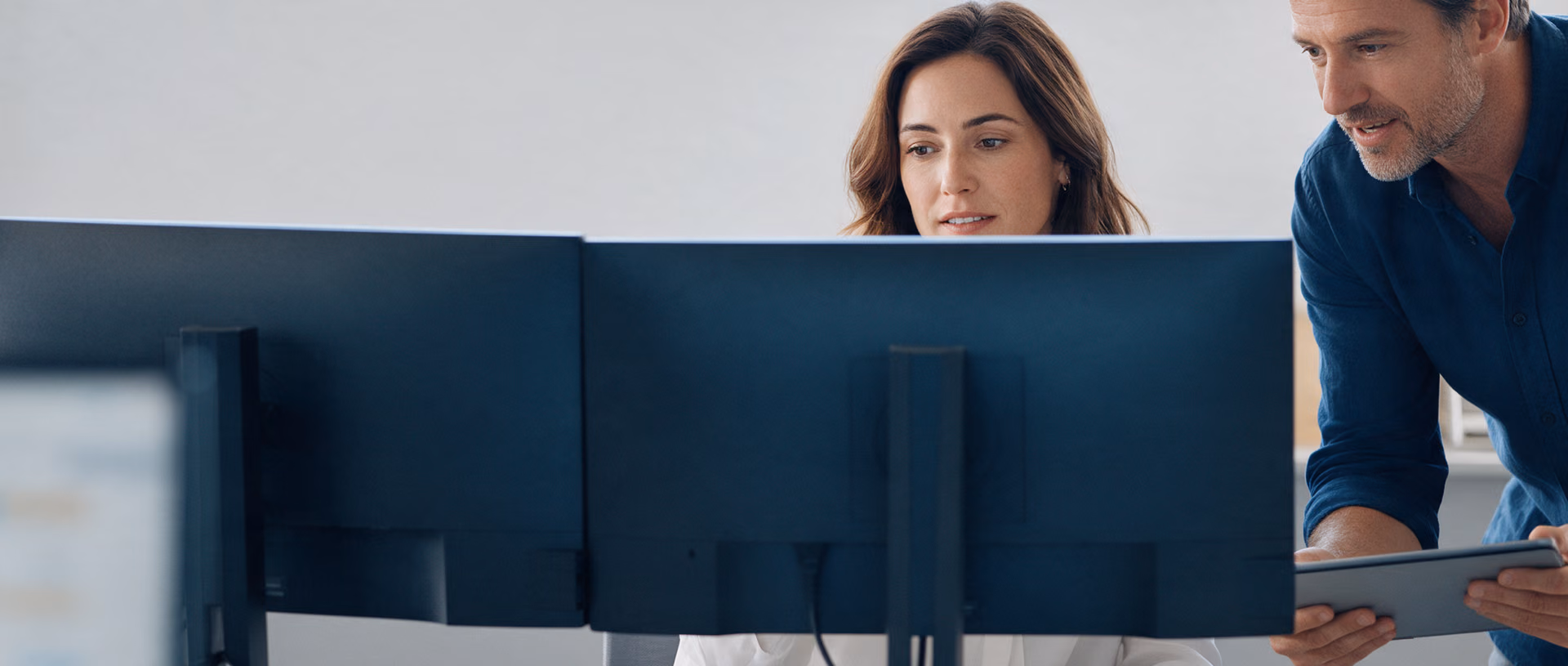 Two colleagues look at a pair of monitors seen from behind at a finance desk, one seated and one standing with a tablet.