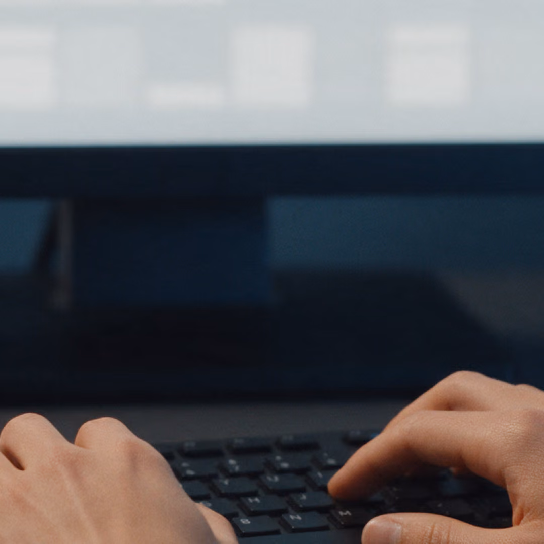 Two hands type on a dark keyboard in front of a monitor showing a blurred grid.