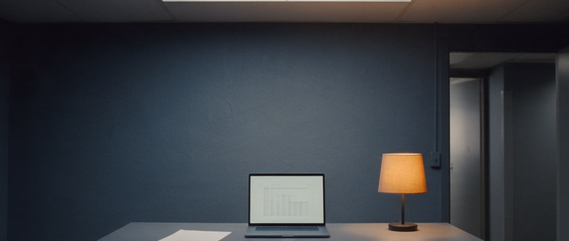 An empty office after dark with a laptop, a lit desk lamp and a single sheet of paper on a dark desk in front of a dark wall.