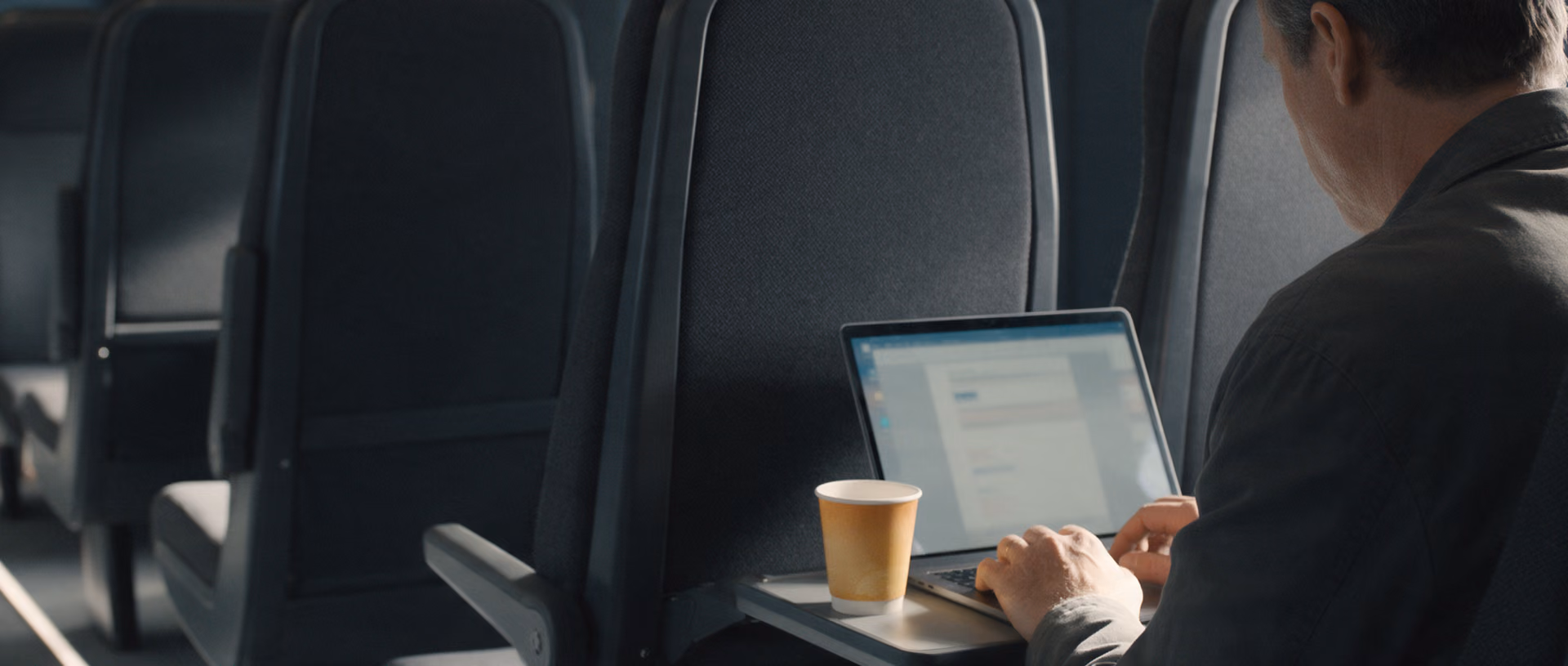 A man seen from behind works on a laptop at a train window seat, a paper cup beside it on the tray table and rows of dark grey seats ahead.