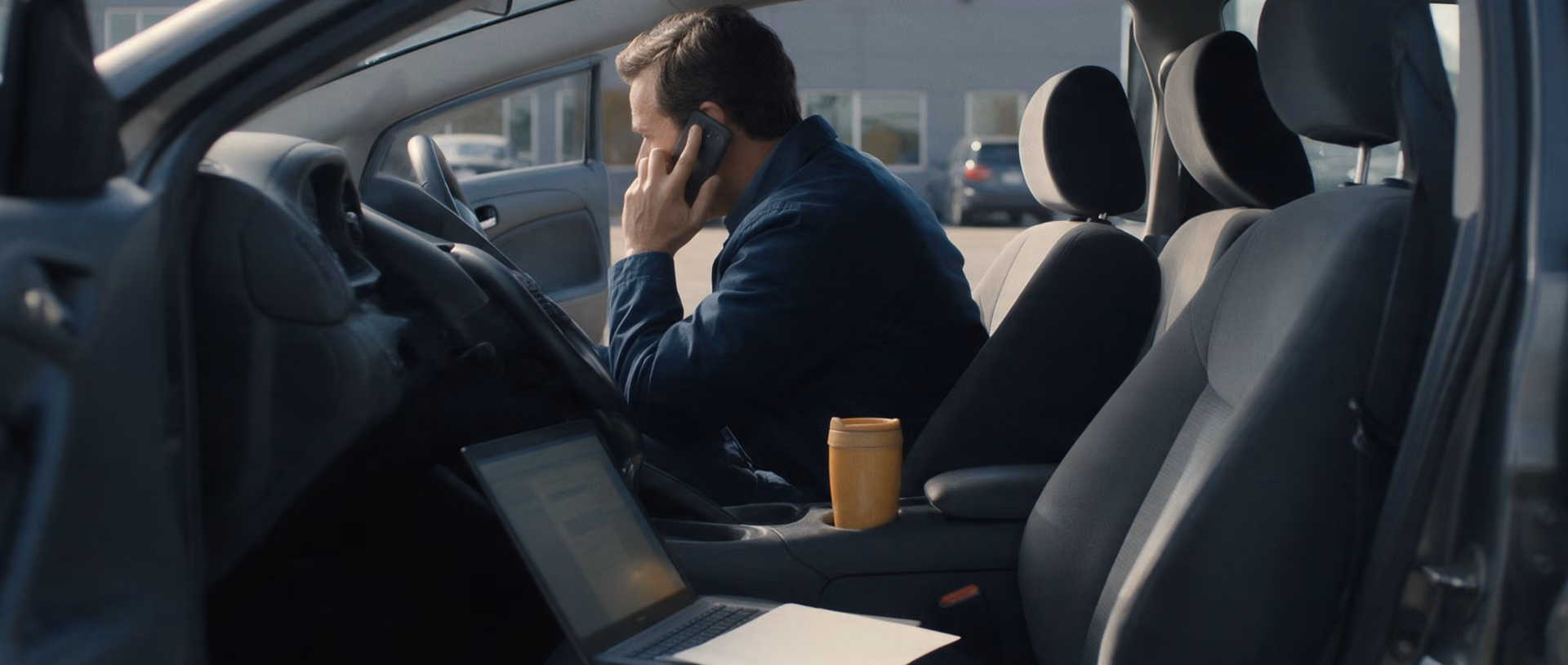 A man sits in the driver seat of a parked car talking on his phone, a laptop open on the passenger seat beside a travel mug.