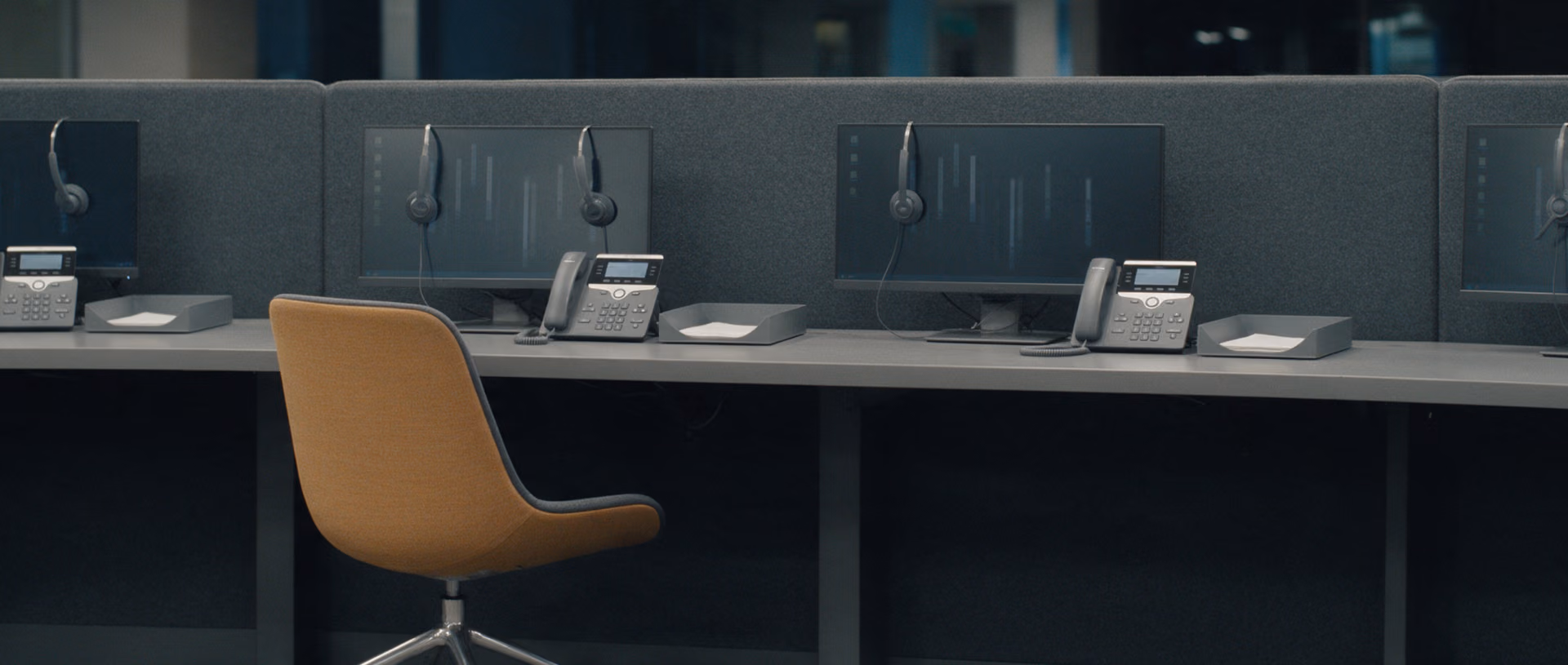 A row of empty telephone workplaces after dark, each with a monitor still on, a headset on its hook and a desk phone, one upholstered chair pushed in.