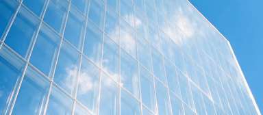 The glass facade of a modern office building reflecting a blue sky with white clouds.