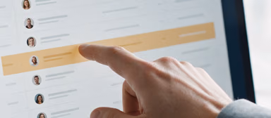 A hand pointing at a contact list in a CRM on a laptop screen, one row highlighted in amber.