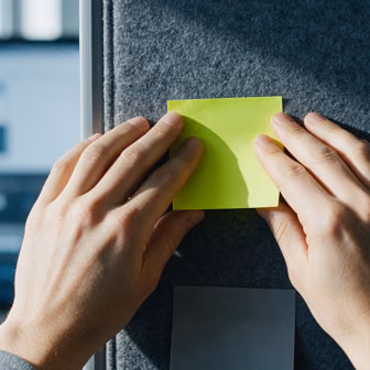 Two hands pressing a lime-yellow sticky note onto a dark grey felt pinboard.