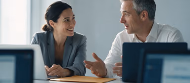 Two colleagues in a bright meeting room turning towards each other in conversation, an amber-sand folder on the table in front of them.