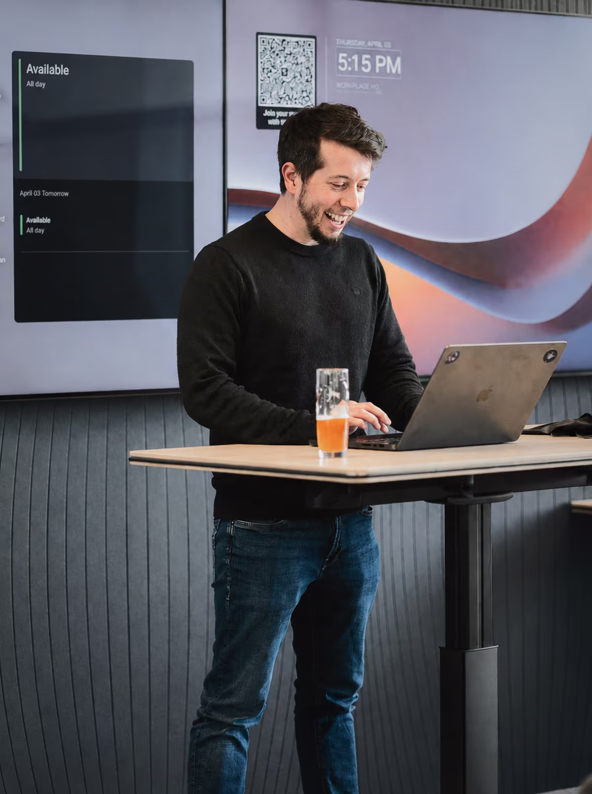 A colleague working at a laptop at a standing desk, with room displays on the wall behind.