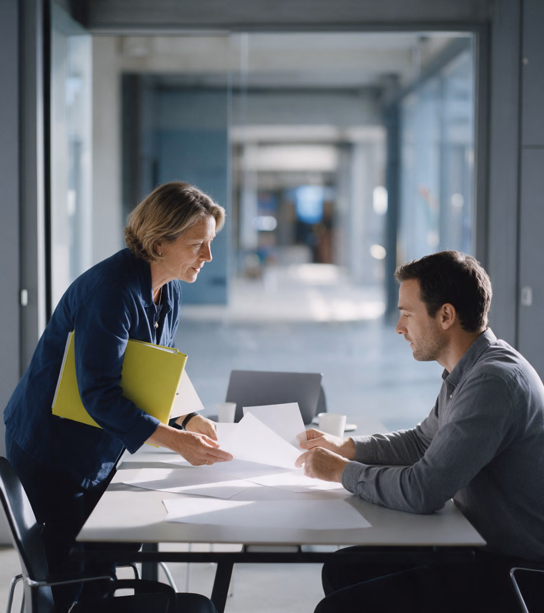 A standing woman with a lime-yellow ring binder under her arm going through printed sheets with a seated colleague in a meeting room whose glass wall looks onto a blurred hall.